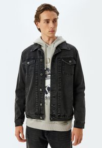 Chaqueta de denim negra con un diseño clásico, que presenta un cierre frontal con botones, dos bolsillos en el pecho y un ajuste holgado. Lleva puesta una sudadera gris debajo.