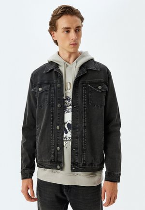 Chaqueta de denim negra con un diseño clásico, que presenta un cierre frontal con botones, dos bolsillos en el pecho y un ajuste holgado. Lleva puesta una sudadera gris debajo.