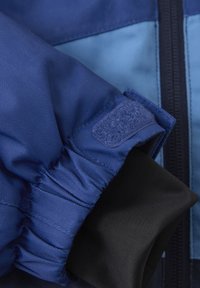 Giacca blu con una superficie esterna testurizzata a motivo chevron, polsini elastici e un morbido rivestimento interno nero. Dettaglio di chiusura in Velcro.
