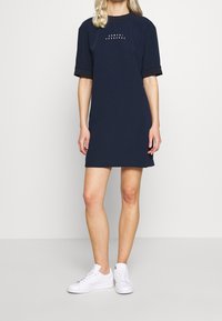 Marinblå bomullsklänning med korta ärmar och rund halsringning. Har ett tryckt mönster på bröstet och svarta muddars. Bärs med vita sneakers.