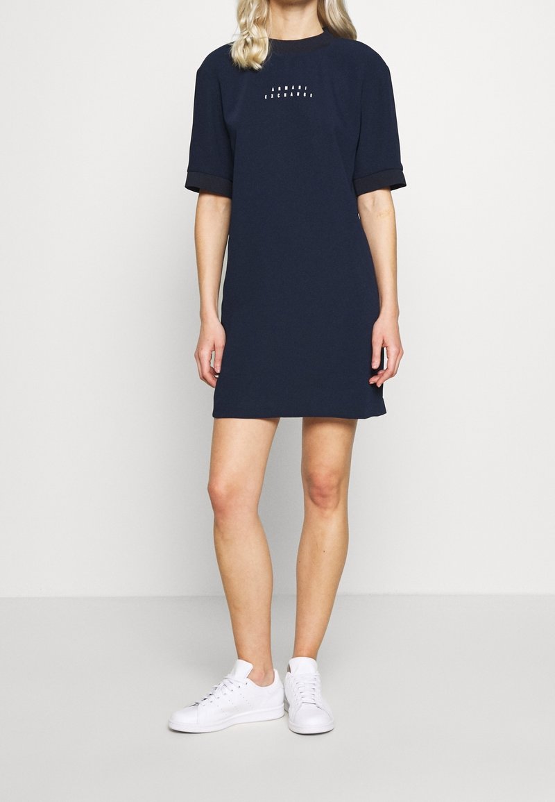 Marinblå bomullsklänning med korta ärmar och rund halsringning. Har ett tryckt mönster på bröstet och svarta muddars. Bärs med vita sneakers.