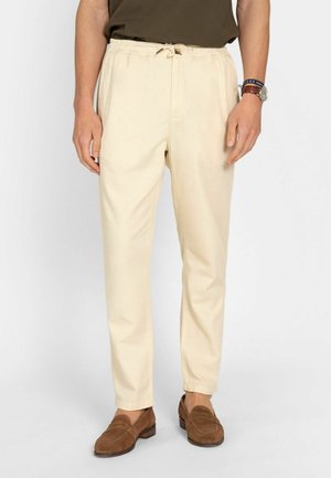 Hombre que lleva pantalones beige con cordón y puños elásticos, mocasines marrones, una camisa oscura y un reloj de pulsera sobre un fondo blanco.