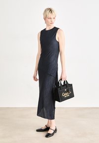 Tummansininen, hihaton mekko, jossa on teksturoitu muotoilu ja epäsymmetrinen helma. Musta käsilaukku, jossa on kultainen "Coach New York" -logo ja yläkahvat.
