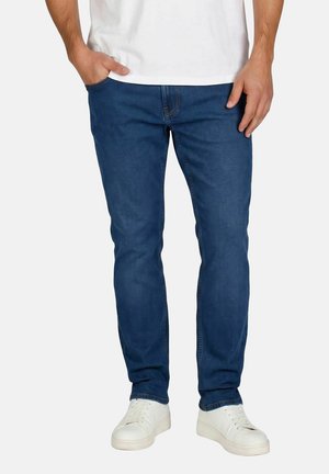 Homme portant un jean bleu, des baskets blanches et un t-shirt blanc, debout avec une main dans la poche devant un fond uni.