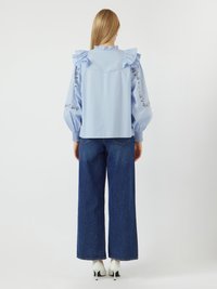 Blusa azzurro chiaro a maniche lunghe con spalle a balze e ricami floreali sulle maniche, abbinata a jeans blu a gamba larga e tacchi bianchi.