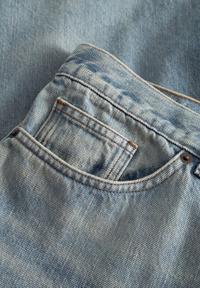 Hellblaue Jeans aus Denim mit einer Taschenapplikation, kontrastierenden braunen Nähten und glatter Textur. Ein dezentes verwaschenes Muster ist erkennbar.