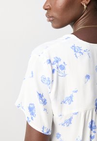 Blusa bianca con stampa floreale blu, maniche corte, scollo rotondo e dettaglio arricciato nella cucitura posteriore. Tessuto morbido e leggero.