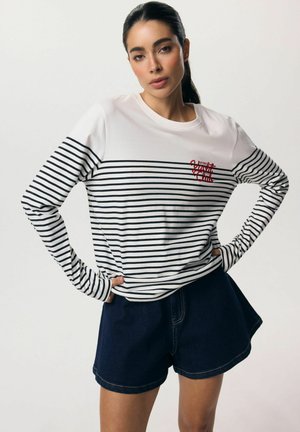 Kobieta z ciemnymi włosami związanymi w kucyk, ubrana w białą i czarną koszulkę w paski z długim rękawem oraz ciemnoniebieskie dżinsowe szorty, ręce na biodrach.