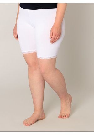 Persoon draagt witte knielange shorts met kanten rand, staand op blote voeten op een eenvoudige neutrale achtergrond.