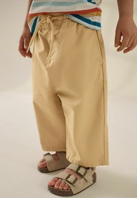 Khaki-kleurige peutershorts met een elastische tailleband en trekkoord. Gemaakt in combinatie met beige sandalen met twee verstelbare gespen.