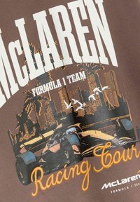 Hnědé tričko s logem týmu McLaren Formula 1, grafikou auta, stylizovaným městským horizontem, palmami a vyšívaným nápisem "Racing Court" v oranžové barvě.