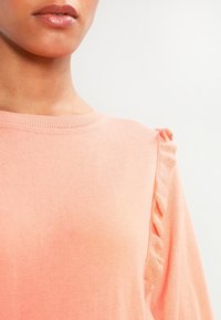 Dlouhý rukáv v broskvové barvě, vyrobený z měkké látky, s volánkovými ozdobami na rameni a klasickým kulatým výstřihem.