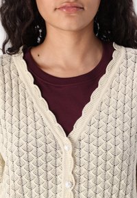 Knit cardigan in cream with scalloped edges, featuring a diamond pattern and three translucent buttons, layered over a maroon shirt.