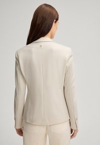 Jasnobeżowa marynarka z delikatnym wzorem w kratkę, o strukturalnym kroju, z zapięciem na jeden guzik i długimi rękawami. Gładka faktura materiału.