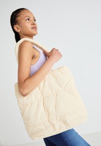 Sac fourre-tout beige moelleux avec texture matelassée, tissu doux et poignées robustes. Le modèle le tient tout en portant un haut lavande et un jean en denim.