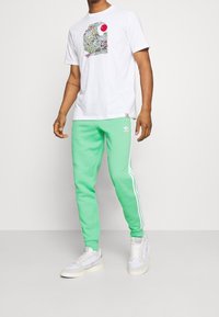 Muž v bílém grafickém tričku, mentolově zelených teplácích Adidas se bílými pruhy a bílých teniskách, stojící na neutrálním pozadí.
