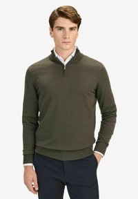 Olivgrön stickad pullover med dragkedja i nacken, ribbade ärmslut och nederkant. Bärs över en vit skjorta med krage, figurnära design, strukturerad tyg.