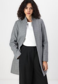 Grijze wolblend jas met een hoge kraag, open voorkant en zijzakken, gecombineerd met zwarte wijde broek. Gladde afwerking, op maat gemaakte pasvorm.