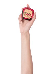 L'Oréal Infallible 24H Fresh Wear foundation i en puderkompakt, med ett genomskinligt röd och svart etui, hålls av en hand mot en ljus bakgrund.