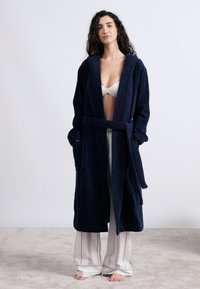 Μπλε ναυτικό μπουρνούζι από πετσέτα, με μακριά μανίκια, δύο μπροστινές τσέπες και ζώνη στη μέση. Φορεμένο πάνω από ανοιχτόχρωμο, ριγέ παντελόνι πιτζάμας.