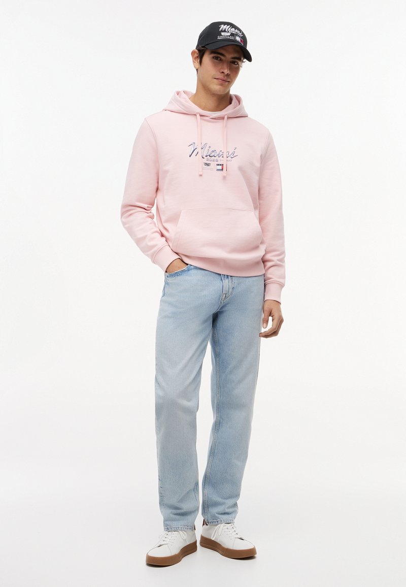 Giovane uomo con cappellino nero, felpa rosa chiaro con scritta "Miami", jeans azzurri e sneaker bianche, in piedi con una mano in tasca.