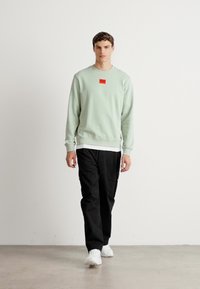 Giovane uomo che cammina con pantaloni neri, sneakers bianche e una felpa verde chiaro con un logo quadrato rosso sul petto, sfondo bianco.