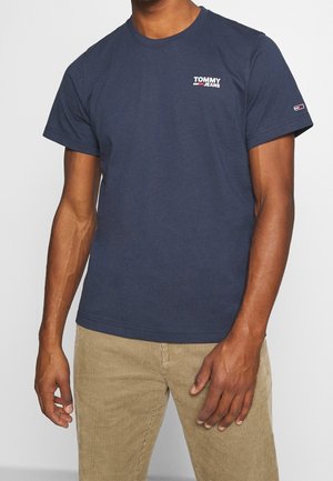 Granatowy bawełniany t-shirt z krótkimi rękawami, z okrągłym dekoltem, logo "TOMMY JEANS" w białym i czerwonym kolorze na piersi, połączony z beżowymi spodniami z aksamitu.