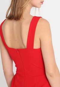 Abito rosso con spalline larghe, corpino aderente e schiena profonda. Presenta una cerniera discreta sul retro ed è realizzato in tessuto morbido.