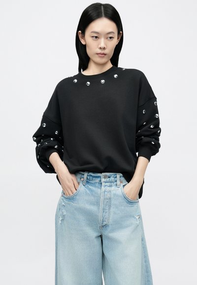 Čierna mikina s striebornými cvokmi pozdĺž výstrihu a rukávov, kombinovaná so svetlomodrými roztrhanými širokými džínsami.