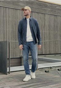 Mörkblå knapp-down skjorta, vit t-shirt, blå jeans och vita sneakers. Man som bär en beige keps står på en träveranda med modern bakgrund.