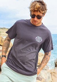 T-shirt en coton gris foncé à manches courtes, avec un motif floral en violet clair sur la poitrine gauche. Porté avec un short vert menthe.