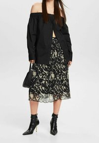 Camisa negra off-shoulder con cuello fruncido, combinada con una falda plisada de patrón negro y crema. Botines negros con punta afilada.