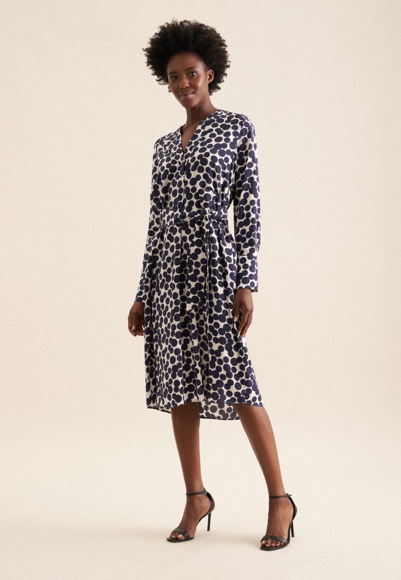 Langärmliges Wickelkleid im Navy- und Weiß-Punkte-Muster mit V-Ausschnitt, tailliertem Bund und fließendem Rock, kombiniert mit Sandalen.