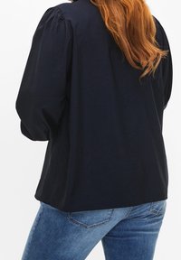 Blusa a maniche lunghe blu navy con spalle puffate e vestibilità rilassata, abbinata a jeans in denim chiaro, con tessuto liscio e strutturato.