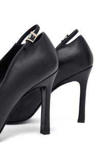 Zapatos de tacón alto de cuero negro con tacones finos y correas para el tobillo que presentan pequeñas hebillas de plata, fotografiados sobre un fondo blanco.