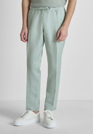 Homme portant un pantalon vert clair à cordon de serrage et des baskets blanches, debout devant un fond uni gris clair.