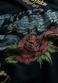 Tessuto nero con uno scorpione blu e una rosa rossa adornati da strass. Il design include foglie verdi e testo dorato.