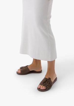 Stopy w brązowych sandałach na płaskiej podeszwie z otwartymi palcami i dużymi kwiatowymi zdobieniami, w połączeniu z białą spódnicą do kostek.