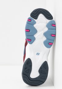 Suela de una zapatilla con banda de rodadura de goma blanca, azul, negra y rosa, con ranuras curvadas y un logo en forma de "S" en el centro.