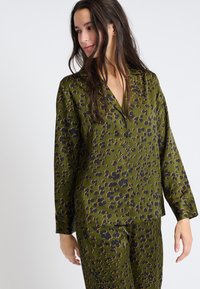 Groen pyjama-set van gladde stof, met een zwart en bruin bloemenpatroon, bestaande uit een knoopsluiting shirt en loszittende broek.