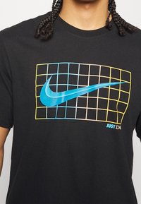 Camiseta de algodón negra con un diseño gráfico que presenta un logo azul de Nike y un patrón en cuadrícula en azul, amarillo y naranja, con el texto "JUST DO IT".