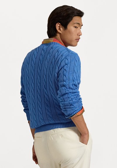 Cable-knit blue sweater with ribbed hem and cuffs, worn over a multi-colored collared shirt, paired with cream-colored pants.