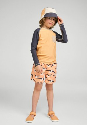 Dziecko w kapeluszu typu bucket z blokami kolorów, żółto-szarym rashguardzie z długim rękawem, brzoskwiniowych szortach z wzorem ptaków oraz pomarańczowych butach do wody.