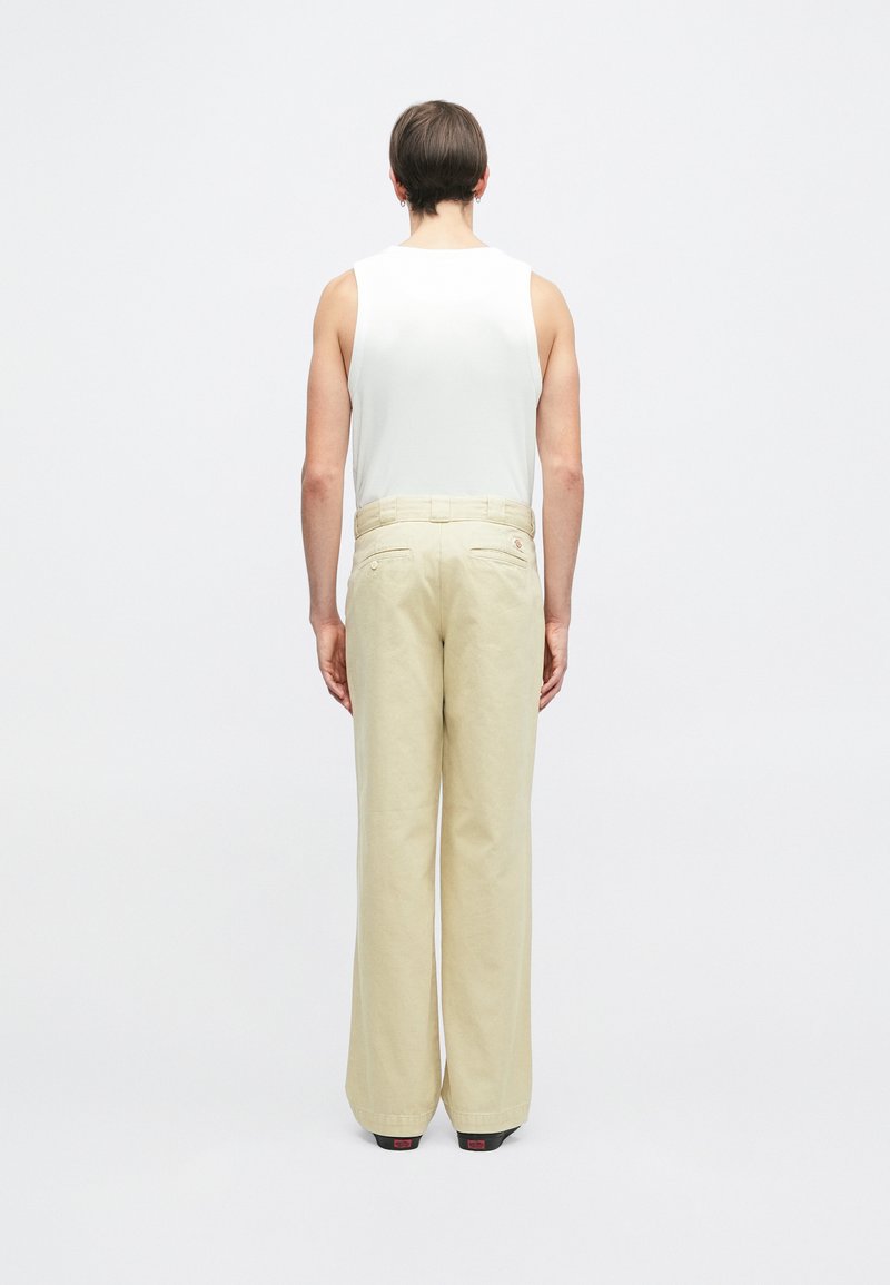 Pantaloni wide-leg beige chiaro con una texture liscia, dotati di due tasche posteriori e dettagli con passanti per cintura, abbinati a un top aderente bianco.