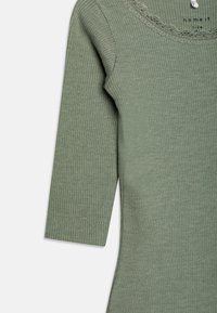 Grønn ribbestrikket topp med trekvartslange ermer og scallop halslinje. Stoffet ser mykt ut med en teksturert overflate og ren hem.