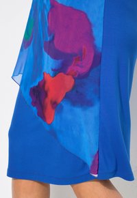 Modré šaty s hladkým materiálem a poloprůhledným overlayem. Overlay obsahuje barevné abstraktní tvary v červené a fialové barvě.