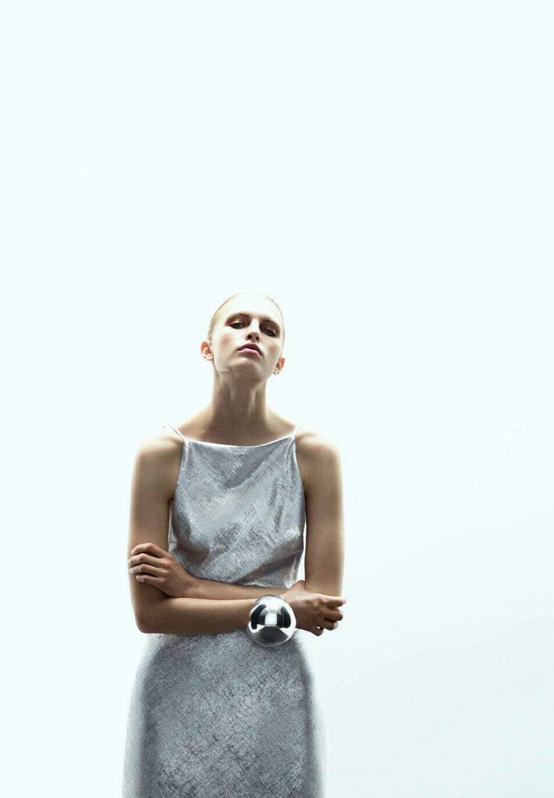 Vestido plateado texturizado con un diseño sin mangas, combinado con una gran pulsera metálica. El fondo es liso y de color claro.