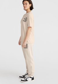 Beige t-shirt met zwarte tekst en loszittende beige trainingsbroek. Witte en zwarte sneakers met zwarte accenten, ontworpen voor comfort.