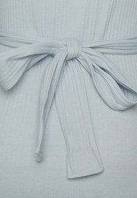 Tecido canelado azul claro com um detalhe de cintura atada. A fita tem uma textura suave, acentuando o design com um laço na frente.