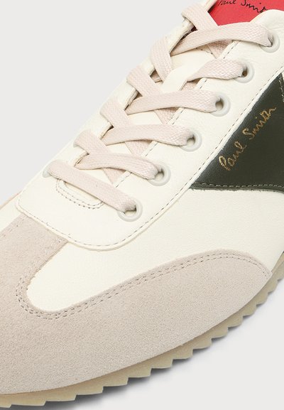 Baskets blanches et beiges avec lacets beiges, bout en daim, panneau latéral en cuir noir avec logo Paul Smith doré, et étiquette rouge sur la languette.